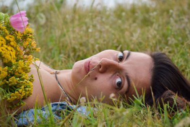 Bir buket kır çiçeğiyle çimlerin üzerinde uzanan çok kültürlü genç bir kadın. Romantizm, şefkat ve kadınlık hissi yayıyor. Büyüleyici bir sahne yaratıyor.