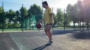 Coşkulu, çok ırklı genç bir kadın, halk parkının spor sahasının ışıl ışıl ışığında bir basketbol topunu güvenle tutuyor. Uygulamasını güneş doğumu egzersizinin canlandırıcı enerjisiyle birleştiriyor.