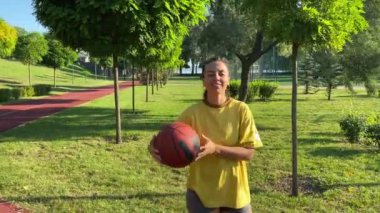 Şafak vakti halka açık parkta poz veren çok ırklı genç bir kadın. Kendine güvenen bir gülüşle zıplayan kadın sporcu topu enerji kararlılığı yayar. Arka planda spor salonu