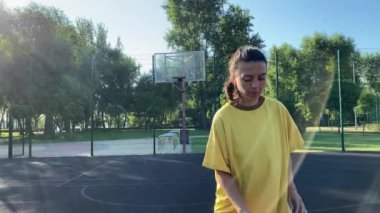 Coşkulu, çok ırklı genç bir kadın, halk parkının spor sahasının ışıl ışıl ışığında bir basketbol topunu güvenle tutuyor. Uygulamasını güneş doğumu egzersizinin canlandırıcı enerjisiyle birleştiriyor.