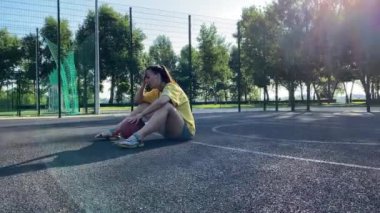 Çok ırklı genç bir kadın elinde basketbol topuyla halka açık park spor sahasında güneşin tadını çıkarıyor. Düşüncelere dalmış, belki de bu güneşli günde arkadaş ya da dostça bir maç için can atıyor.