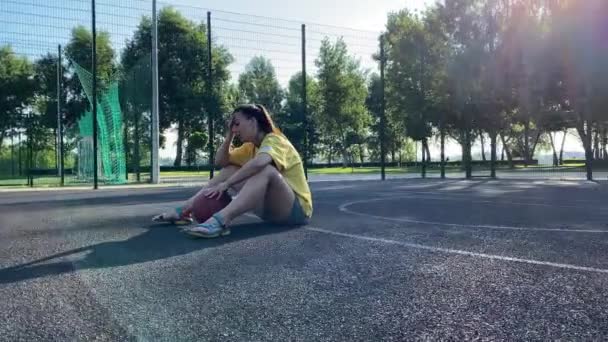 多种族的年轻女子坐在篮球场上 在晨阳下沐浴在公园的运动场上 在这个阳光灿烂的日子里 她发现自己陷入了沉思 也许是渴望有人陪 或是渴望一场友爱的比赛 — 图库视频影像