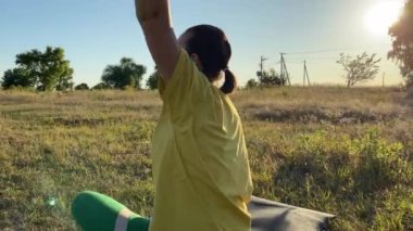 Canlı spor kıyafetleri içindeki genç bir kadın, altın güneş ışığı ve açık mavi gökyüzünde hareketli bir spor rutinine hazırlanarak, sakin bir kırsal alanda hareketli bir ısınma egzersizine girişiyor.