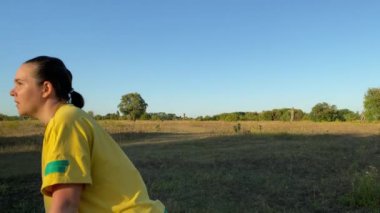 Canlı spor kıyafetleri içindeki genç bir kadın, altın güneş ışığı ve açık mavi gökyüzünde hareketli bir spor rutinine hazırlanarak, sakin bir kırsal alanda hareketli bir ısınma egzersizine girişiyor.