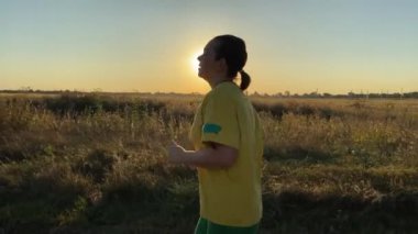Canlı spor kıyafetleri içinde genç bir kadın manzaralı kırsal manzarayla çevrili sakin köy yolunda koşuyor, altın akşam güneşiyle yıkanmış. Egzersiz rutini, aktif yaşam tarzı
