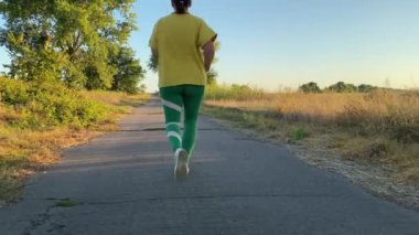 Canlı spor kıyafetleri içinde genç bir kadın, manzaralı kırsal manzarayla çevrili sakin kır yolu boyunca koşuyor, altın akşam güneşinin altında yıkanıyor. Egzersiz rutini, aktif yaşam tarzı