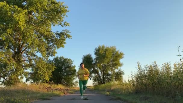 若い女性は穏やかな田舎の道路に沿って夕方のジョギングを活性化することに従事しています 低い角度のビュー 女性のジョギングは 夕日の黄金の時間に美しい田舎の風景で彼女の息をキャッチするために一時停止 — ストック動画