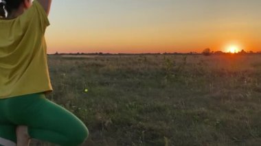 Yoga ağacında dengede duran sakin genç bir kadın kırsal alanda altın saatinde poz veriyor. Güneş arka planda batıyor, sıcak ve davetkar bir ortam yaratıyor. Kadın kararlı, pozu zarif.