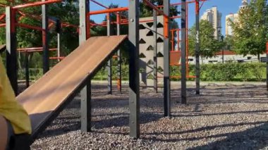 Halka açık bir parktaki açık hava spor salonunda, sabah güneşinin altında dambılla kol pompalama egzersizi yapan genç bir kadın. Oyun alanı ekipmanları üzerinde güç eğitimi veriliyor