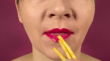 Renkli dudakları olan tanınmayan genç bir kadın yemek yiyor, lezzetli yemekler yiyor ve her ısırığı içeride çiğniyor. Kapalı ağızlı pembe ruj mor arka planda izole edilmiş kızarmış patateslerin tadını çıkarıyor.