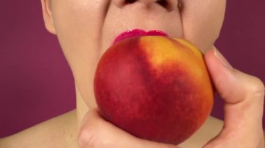 Renkli dudakları olan tanınmayan genç bir kadın olgun şeftali yer ve içerideki sulu meyveyi ısırır. Kapalı ağızlı pembe ruj mor arka planda izole edilmiş tatlı yemeğin tadını çıkarıyor.