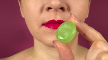 Rengarenk dudakları olan tanınmayan genç bir kadın tatlı yiyecekler yiyor, lezzetli lezzet emici sert şekerlerin tadını çıkarıyor. Kapalı ağız pembe ruj emme lolipop emme mor arka planda izole