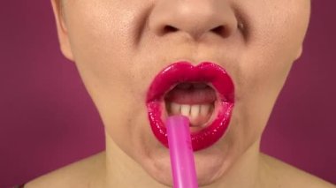 Renkli dudakları olan tanınmayan genç bir kadın pembe bir pipetle alkollü bir içecek yudumlayarak mor arka planda kapalı alanda göz alıcı bir yakın çekim yaratıyor.