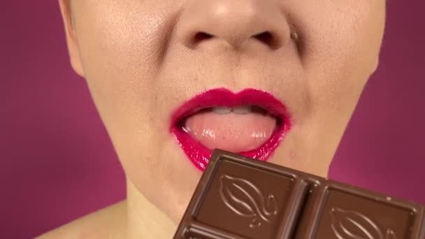 鮮やかな色の唇を持つ認識できない若い大人の女性は 甘い食べ物 おいしいチョコレートを食べ 屋内で各噛み合う パープルで隔離されたクローズアップ口ピンクの口紅の香りのデザート — ストック動画