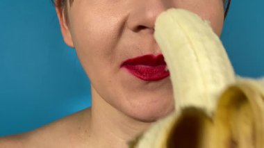 Canlı renkli dudakları olan tanınmayan genç bir kadın baştan çıkarıcı bir şekilde ısırıyor ve içeride taze muz yalıyor. Kapalı dudaklı kırmızı ruj mavi arka planda izole edilmiş tatlı meyvenin tadını çıkarıyor