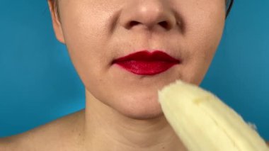 Renkli dudakları olan tanınmayan genç bir kadın olgun bir muzu yer ve içerideki meyveleri ısırır. Kapalı dudaklı kırmızı ruj mavi arka planda izole edilmiş tatlı yemeğin tadını çıkarıyor.