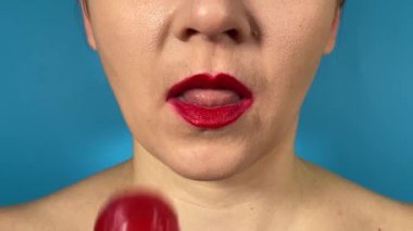 Renkli dudakları olan tanınmayan genç bir kadın tatlı yiyecekler yiyor, evde lolipop yalıyor. Kapalı dudaklı kırmızı ruj mavi arka planda izole edilmiş bir çubukta tatlı sert şekerin tadını çıkarıyor.