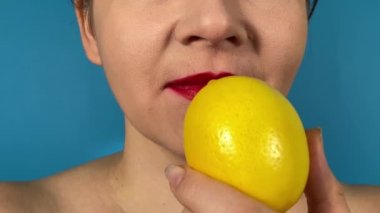 Renkli dudakları olan tanınmayan genç bir kadın olgun limonu ısırıyor, meyveyi içeride yiyor. Kapalı ağız kırmızı ruj. Mavi arka planda taze narenciye tadı.