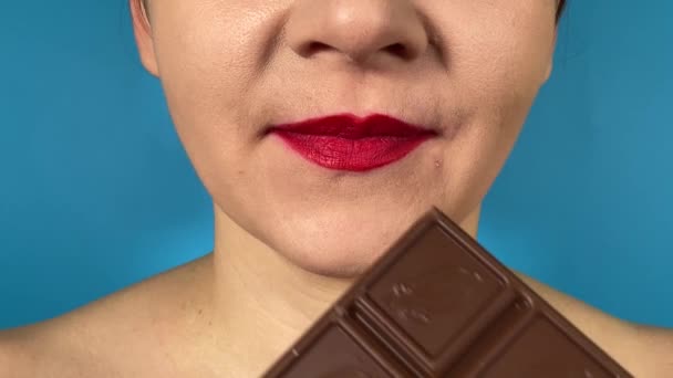 鮮やかな色の唇を持つ認識できない若い大人の女性は 甘い食べ物を食べ 屋内でチョコレートの大きな部分に噛み付きます 青い背景に隔離されたクローズアップ口紅の口紅の香りのデザート — ストック動画
