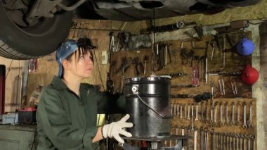 Tulum giymiş genç bir kadın bir araba bakım istasyonunda çalışıyor, kaldırılmış bir araca teknik bakım yapıyor, eski motor yağını boşaltıyor ve değiştiriyor. Arkaplanda çeşitli araçlar