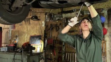 Tulumlu genç bir kadın bir araba servis istasyonunda çalışıyor, kapalı alanda teknik araba bakımı yapıyor. Yükseltilmiş bir aracın yağ filtresini değiştiren kadın tamirci. Arka planda duran çeşitli araçlar