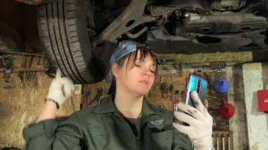 Tulumlu genç bir kadın, kapalı alanda mobil bir uygulama kullanarak bir araba servis istasyonunda canlı yayın yapıyor. Kadın tamirci blogu, internet üzerinden işiyle ilgili görüşlerini paylaşıyor.