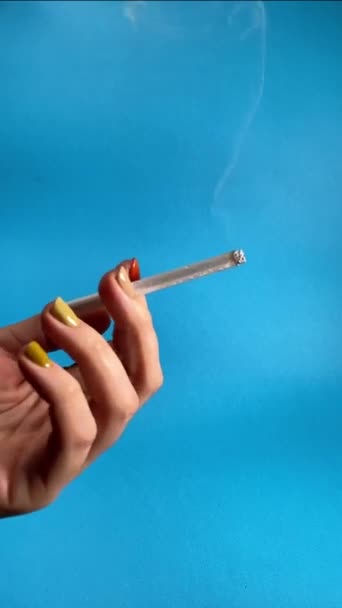 女性的手带着一支吸烟的香烟出现在人们的视野中 用蓝色背景隔离的可见的卷烟来展示吸烟的行为 无法辨认的妇女将烟草产品存放在室内 — 图库视频影像