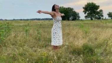 Eğlence, boş zaman, açık hava. Çok ırklı elbiseli çekici bir genç kadın doğanın güzelliğine hayran, çayırlarda geziniyor, yaz mevsiminde çimenlerin ve kır çiçeklerinin tadını çıkarıyor.
