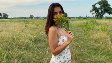 Eğlence, boş zaman, açık hava. Çok ırklı elbiseli çekici bir genç kadın doğanın güzelliğine hayran, çayırlarda geziniyor, yaz mevsiminde çimenlerin ve kır çiçeklerinin tadını çıkarıyor.