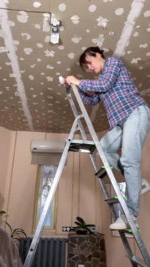 Evde tadilat yapan bir kadın, evin arka planında tadilat yapıyor. Dikey çekim, DIY konsepti. Tamirci kadın ampulü lambayla değiştirip elektrik lambasını tamir ediyor.