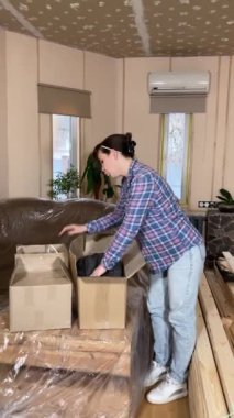 Güzel, genç bir kadın evin arka planında inşaat malzemelerinin olduğu kutuları taşıyarak taşınıyor. Dikey çekim, DIY konsepti. Becerikli kadın, yenileme hazırlıkları için ev geliştirme çalışmalarına başladı.