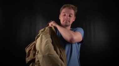 Şık bir genç adam, günlük görünüşü olan, sıradan bir kirli sakalı olan mavi bir tişört giyer, keskin bakışları, asgari siyah arkaplana karşı kendine güveni tam bir tutum sergiler.