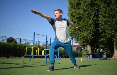 Odaklanmış genç bir atlet dinamik bir şekilde ısınma egzersizleri yapıyor ve dışarıda esniyor. Atletizm kıyafetlerini giyip titizlikle vücudu düzenli bir rutine göre fiziksel taleplere hazırlıyor.