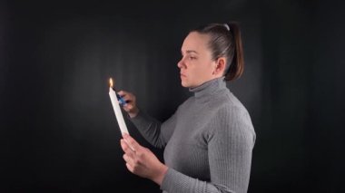 Çekici genç bir kadın, alevini sessiz bir tefekkür havasıyla söndürmeden önce mum fitili yakar. Simsiyah arka planda basit bir ayin farkındalığı hissi. Düşünceli bir yaşam tarzı