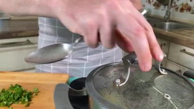 Önlüklü genç bir adam ev mutfağında yemek pişiriyor ve bir tencere kaynar suya malzeme ekleyerek çorba hazırlıyor. İçerde sebze ve baharatları birleştiriyor. Ev yemeği ve aşçılık tutkusu