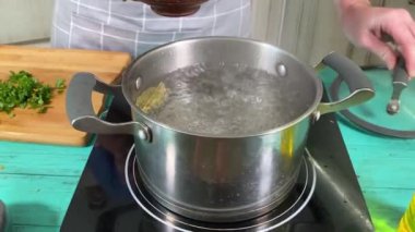 Önlüklü genç bir adam ev mutfağında yemek pişiriyor ve bir tencere kaynar suya malzeme ekleyerek çorba hazırlıyor. İçerde sebze ve baharatları birleştiriyor. Ev yemeği ve aşçılık tutkusu