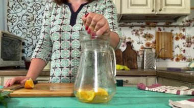 Önlüklü genç bir kadın ev mutfağında ferahlatıcı limonata yapmak için malzeme hazırlıyor. Narenciye, nane ve buzu birleştirip serin, canlandırıcı bir içecek yapıyor. Ev yapımı yaz içecekleri.