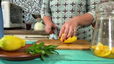 Önlüklü genç bir kadın ev mutfağında ferahlatıcı limonata yapmak için malzeme hazırlıyor. Narenciye, nane ve buzu birleştirip serin, canlandırıcı bir içecek yapıyor. Ev yapımı yaz içecekleri.