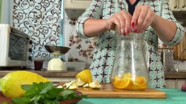 Önlüklü genç bir kadın ev mutfağında ferahlatıcı limonata yapmak için malzeme hazırlıyor. Narenciye, nane ve buzu birleştirip serin, canlandırıcı bir içecek yapıyor. Ev yapımı yaz içecekleri.