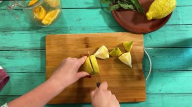 Önlüklü genç bir kadın ev mutfağında ferahlatıcı limonata yapmak için malzeme hazırlıyor. Narenciye, nane ve buzu birleştirip serin, canlandırıcı bir içecek yapıyor. Ev yapımı yaz içecekleri.