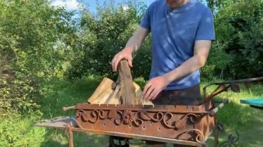 Beyaz bir adam dışarıda yemek pişirmek için ızgara hazırlıyor, güçlü bir ateş yakmak için odun hazırlıyor. Sıcak bir arka bahçeye kurulur, barbekü hazırlıkları yapılır, açık hava eğlencesi ve mutfak ile harmanlanır.