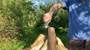 Beyaz bir adam dışarıda yemek pişirmek için ızgara hazırlıyor, güçlü bir ateş yakmak için odun hazırlıyor. Sıcak bir arka bahçeye kurulur, barbekü hazırlıkları yapılır, açık hava eğlencesi ve mutfak ile harmanlanır.