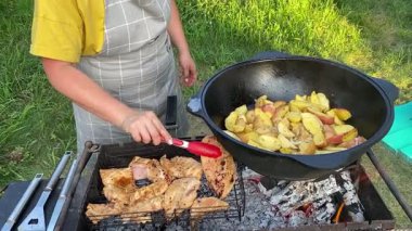 Önlüklü güzel bir kadın açık havada yemek hazırlıyor. Izgarada ateş yakıyor. Rahat bir açık hava ortamında malzemelerle ustalıkla uğraşırken sıcak havanın tadını çıkarıyor..