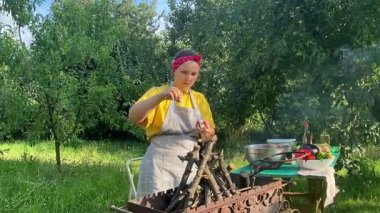 Önlüklü güzel bir kadın açık havada yemek hazırlıyor. Izgarada ateş yakıyor. Rahat bir açık hava ortamında malzemelerle ustalıkla uğraşırken sıcak havanın tadını çıkarıyor..