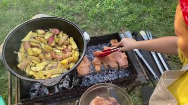 Önlüklü güzel bir kadın açık havada yemek hazırlıyor. Izgarada ateş yakıyor. Rahat bir açık hava ortamında malzemelerle ustalıkla uğraşırken sıcak havanın tadını çıkarıyor..