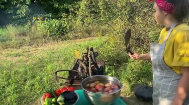 Önlüklü güzel bir kadın açık havada yemek hazırlıyor. Izgarada ateş yakıyor. Rahat bir açık hava ortamında malzemelerle ustalıkla uğraşırken sıcak havanın tadını çıkarıyor..