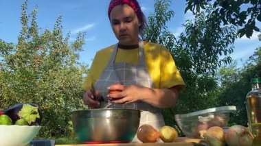 Önlüklü güzel bir kadın açık havada yemek hazırlıyor. Izgarada ateş yakıyor. Rahat bir açık hava ortamında malzemelerle ustalıkla uğraşırken sıcak havanın tadını çıkarıyor..
