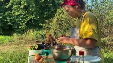 Önlüklü güzel bir kadın açık havada yemek hazırlıyor. Izgarada ateş yakıyor. Rahat bir açık hava ortamında malzemelerle ustalıkla uğraşırken sıcak havanın tadını çıkarıyor..