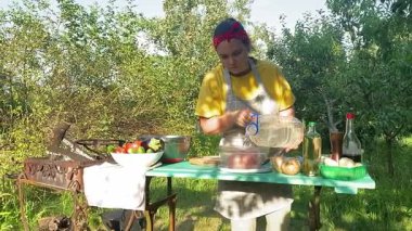 Önlüklü güzel bir kadın açık havada yemek hazırlıyor. Izgarada ateş yakıyor. Rahat bir açık hava ortamında malzemelerle ustalıkla uğraşırken sıcak havanın tadını çıkarıyor..