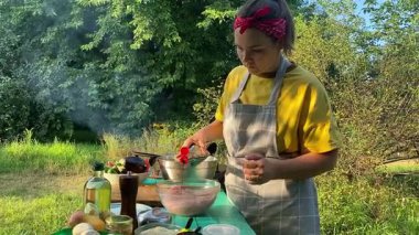 Önlüklü güzel bir kadın açık havada yemek hazırlıyor. Izgarada ateş yakıyor. Rahat bir açık hava ortamında malzemelerle ustalıkla uğraşırken sıcak havanın tadını çıkarıyor..